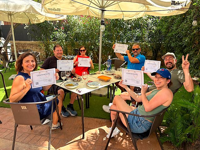 Workshop guests at a table under the umbrellas holding up their paella certificates